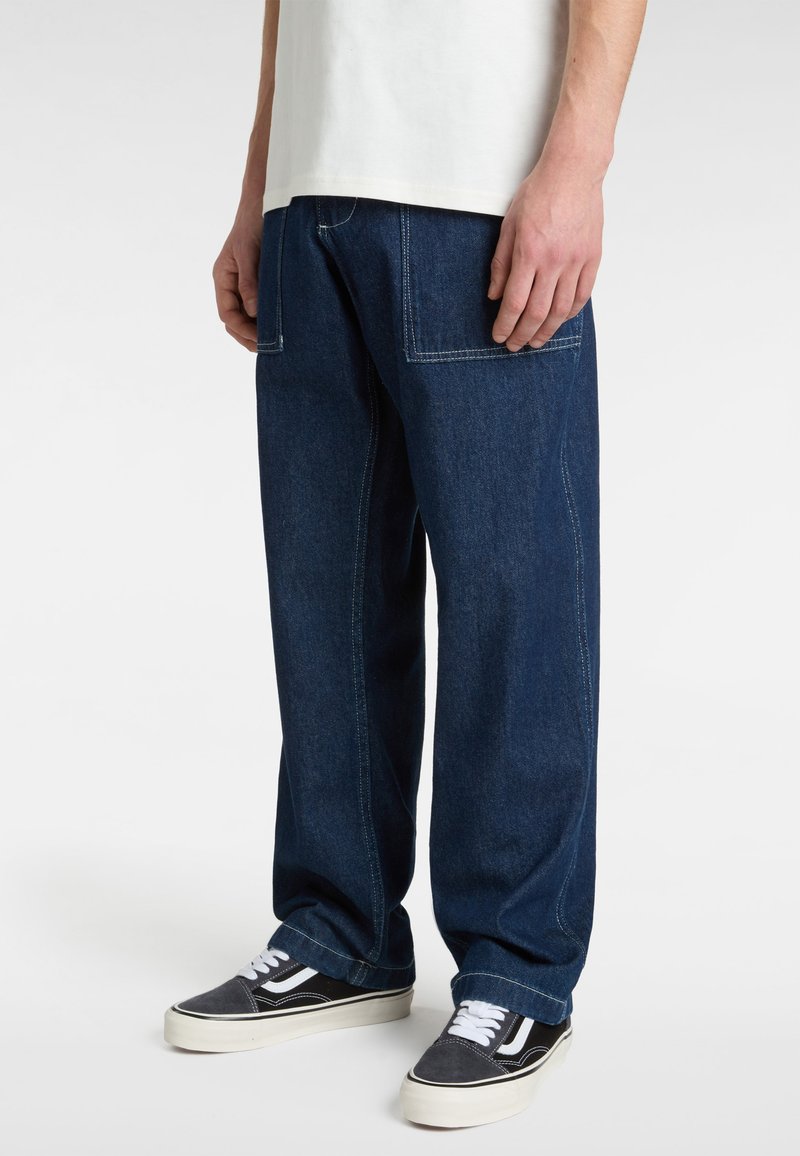 Pantalones de mezclilla anchos en azul oscuro con costuras visibles, dos bolsillos frontales y zapatillas casuales en blanco y negro. El material parece robusto y texturizado.