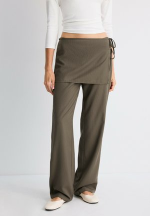 Femme portant un pantalon large à rayures olive avec une superposition de jupe portefeuille attachée et des chaussures plates crème, debout sur fond blanc.