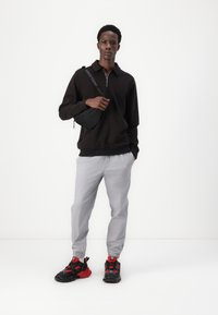 Crna zip-up dukserica, sive tapered hlače i crno/crvene tenisice. Mala crna ušna torba s remenom. Glatke teksture i moderan dizajn.