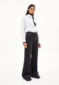 Vit skjorta med knappar, svart polotröja och vida svarta jeans med texturerad yta. Skorna har svarta fransar och pärlor.