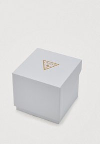 Witte geschenkdoos met een textuur en een gouden driehoekig Guess-logo op het bovenoppervlak, voorzien van een omslaand deksel en vierkante vorm.