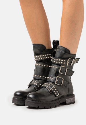 Bottes noires en cuir à hauteur de la cheville avec des clous argentés et plusieurs boucles portées sur des jambes nues, sur un fond uni.