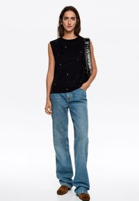 Czarny top bez rękawów z cekinowymi zdobieniami, połączony z jasnoniebieskimi szerokimi spodniami i brązowymi zamszowymi butami. Torba ma błyszczące wykończenie.