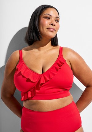 Sebavedomá žena so stredným odtieňom pokožky a čiernymi vlasmi nosi červené dvojdielne plavky so volánmi, pózuje na bielom pozadí.