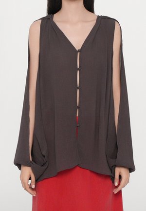 Blusa sin mangas de color gris oscuro con botones de lazo en la parte delantera y tela drapeada sobre los brazos, combinada con una falda roja.