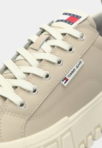Sneaker sintetica beige con lacci bianchi e occhielli argentati. Distintivo tag con il logo "TOMMY JEANS" e suola bianca textured.