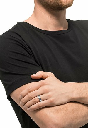 Schwarz kurzärmliges T-Shirt an einer Person mit verschränkten Armen, die einen silbernen und schwarzen zweifarbigen Ring an der linken Hand trägt.