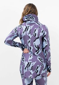 Langärmliges, kapuzenförmiges Oberteil in lila mit blauem und schwarzem Camouflage-Muster. Aus glattem, elastischem Stoff hergestellt. Mit abgerundetem Saum und Daumenlöchern.