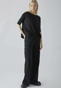 Μαύρο oversized πουκάμισο με φαρδύ ντεκολτέ συνδυασμένο με μαύρο παντελόνι cargo που διαθέτει μεγάλες τσέπες, κατασκευασμένο από μαλακό ύφασμα. Μαύρες επίπεδες παπούτσια.