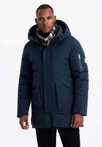 Mörkblå puffjacka med huva, som har dragkedjestängning, framfickor och sidodetaljer. Bärs över en rutig scarf, med en slät textur.