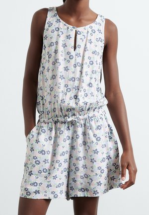 Bezrekawny kombinezon w kwiatowy wzór z niebieskimi i różowymi kwiatami, z elastyczną talią i wiązaniem, noszony przez osobę z rękami w kieszeniach na jednolitym tle.