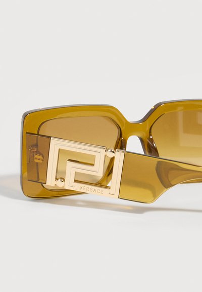 Κίτρινα τετράγωνα υπερμεγέθη γυαλιά ηλίου με χρυσή γεωμετρική λεπτομέρεια λογότυπου Versace στον βραχίονα του σκελετού σε ανοιχτό φόντο.