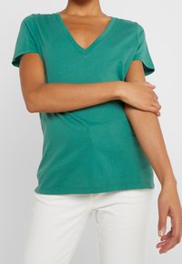 Persona che indossa una maglietta verde a V con maniche corte e pantaloni bianchi, con un braccio incrociato sopra l'altro su uno sfondo neutro.