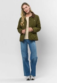 Prešívaná olivovo-zelená bunda s hnedým zamatovým golierom, patentkami a jemným vyšitým detailom, oblečená cez krémový sveter a džíny.