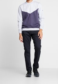 Dlouhý rukáv sportovního trička v světle šedé a tmavě fialové barvě, s kulatým výstřihem a kontrastním trojúhelníkovým vzorem. Doplněno černými kalhotami a botami.