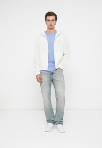 Λευκή ζακέτα με φερμουάρ, ανοιχτό μπλε μπλουζάκι, ανοιχτό τζιν παντελόνι, λευκά αθλητικά παπούτσια. Κανονική εφαρμογή, λεία υφή, χωρίς μοτίβα ή ορατά λογότυπα.