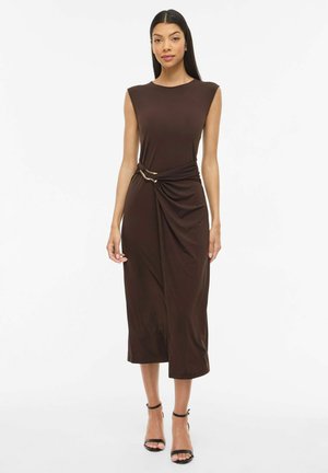 Kvinde iført en ærmeløs, mørkebrun midi-kjole med en rynket taljedetalje, der er sikret med et gyldent bælte med to ringe, kombineret med sorte ankelremssandaler.