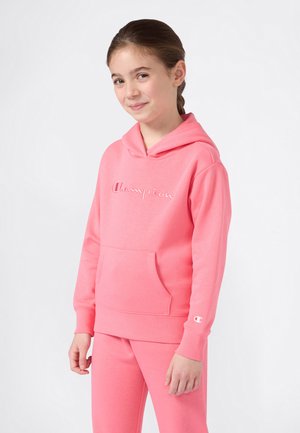 Jeune fille souriante, portant un hoodie Champion rose avec une poche avant et un pantalon rose assorti, se tenant devant un fond blanc uni.