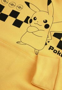 Żółta bluza z grafiką czarnego Pikachu, wzorem w szachownicę i napisem "Pokémon". Rybkowane mankiety i gładka faktura.