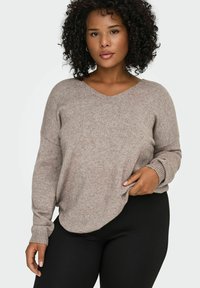 Femme aux cheveux bouclés portant un pull à col en V marron clair et un pantalon noir, posant avec une main tenant l'ourlet du pull.