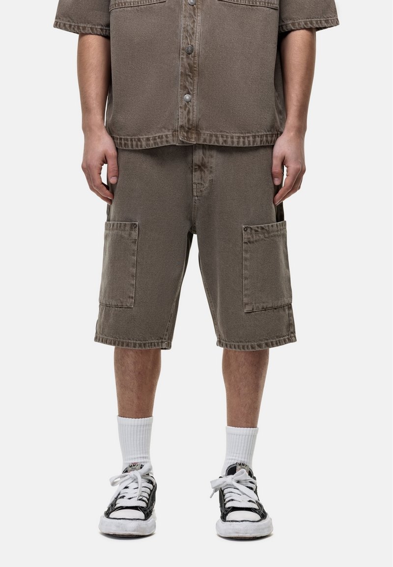 Chemise à manches courtes en denim brun avec des boutons, associée à un short cargo assorti avec deux grandes poches avant, portée avec des chaussettes blanches texturées et des baskets noires.