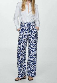 Wijde broek met een blauw-witte spiraalpatroon, gemaakt van een lichte stof. Gecombineerd met een strakke witte blouse en sandalen.