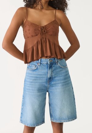 Vrouw draagt een mouwloze bruine top met ruches en kanten details en lichtblauwe knielange denim shorts.