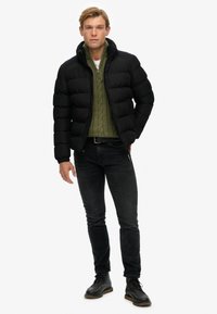 Chaqueta puff negra con cuello alto, usada sobre un suéter de punto de cable verde. Unos jeans oscuros de corte ajustado y botas negras con cordones completan el look.