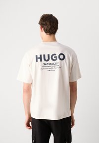Weißes Baumwoll-T-Shirt mit einem großen dunkelsblauen "HUGO"-Logo und Text auf der Rückseite. Kurze Ärmel, Rundhalsausschnitt und lässiger Schnitt.