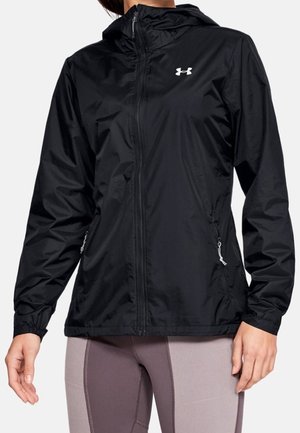 Veste noire à capuche zippée avec des poignets élastiques portée sur des leggings bicolores violets sur une personne debout.