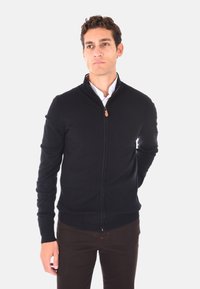 Maglione nero con zip, collo alto, design aderente, polsini e orlo a costine, realizzato in morbido tessuto a maglia. Indossato sopra una camicia bianca.