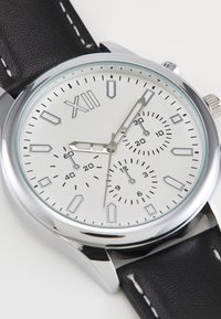 Montre analogique argentée avec un cadran texturé blanc, index noirs, sous-cadrans et un bracelet en cuir noir avec des coutures blanches.