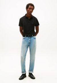 Mladý muž stojící se založenýma rukama za zády, oblečený v černé polokošili, světle modrých džínách, černém opasku a černých botách na bílém pozadí.