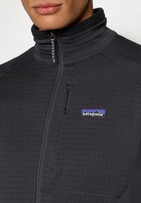 Férfi fekete cipzáros Patagonia dzsekiben, magas gallérral és látható cipzáras mellzsebbel.