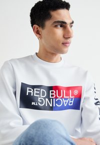 Sudadera blanca con un diseño gráfico audaz que presenta un bloque de colores en rojo, azul y negro, junto con el texto "RED BULL RACING". Incluye cuello de canalé.