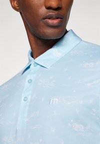 Hellblaues Polohemd mit einem dezenten weißen Muster aus Häusern und Landschaften, ausgestattet mit einem Kragen, drei Knöpfen und einem kleinen gestickten Logo.