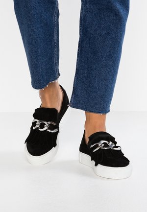 Czarne zamszowe buty wsuwane z frędzlami i srebrnym łańcuszkiem jako akcentem. Białe gumowe podeszwy, zestawione z krótkimi niebieskimi dżinsami.