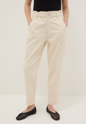 Frau trägt hoch taillierte, elfenbeinfarbene Hose mit Taschen vorne, elastischem Bund und schwarzen, flachen Schuhen mit offener Webart, steht mit den Händen in den Taschen.