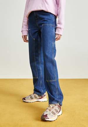 Person iført brede blå jeans med lappede lommer og beige og bordeaux sneakers, stående på en sennepsgul gulv.
