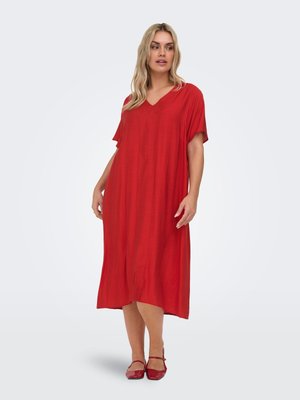 Ženska s svetlimi lasmi, oblečena v široko rdečo midi obleko s kratkimi rokavi ter usklajene rdeče Mary Jane čevlje, stoji na svetli podlagi.