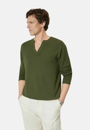 Uomo che indossa una maglia verde oliva a maniche lunghe con scollo a V e pantaloni bianchi, in piedi con le mani nelle tasche, guarda verso sinistra su sfondo semplice.