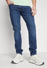Mörkblå denimjeans med slim fit, med traditionell femfickorsdesign. Bärs med vita sportskor och en ljusgrön skjorta.