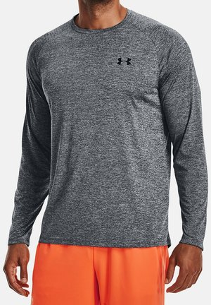 Mann trägt ein eng anliegendes graues langärmeliges Sportshirt mit schwarzem Logo auf der Brust und leuchtend orangefarbene Shorts vor weißem Hintergrund.