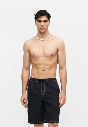 Άνδρας χωρίς μπλούζα με μαύρο μαγιό και λευκό κολιέ από χάντρες στέκεται μπροστά σε λευκό φόντο, κοιτάζοντας ευθεία με ουδέτερη έκφραση.