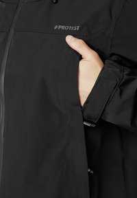 Czarna wodoodporna kurtka zapinana na zamek, z regulowanymi mankietami i logotypem na klatce piersiowej. Posiada boczną kieszeń, w której znajduje się ręka.