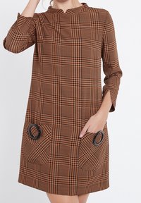 Brun och svart rutig klänning med Mandarin-krage, trekvartsärmar och två cirkulära svarta detaljer på fickorna.