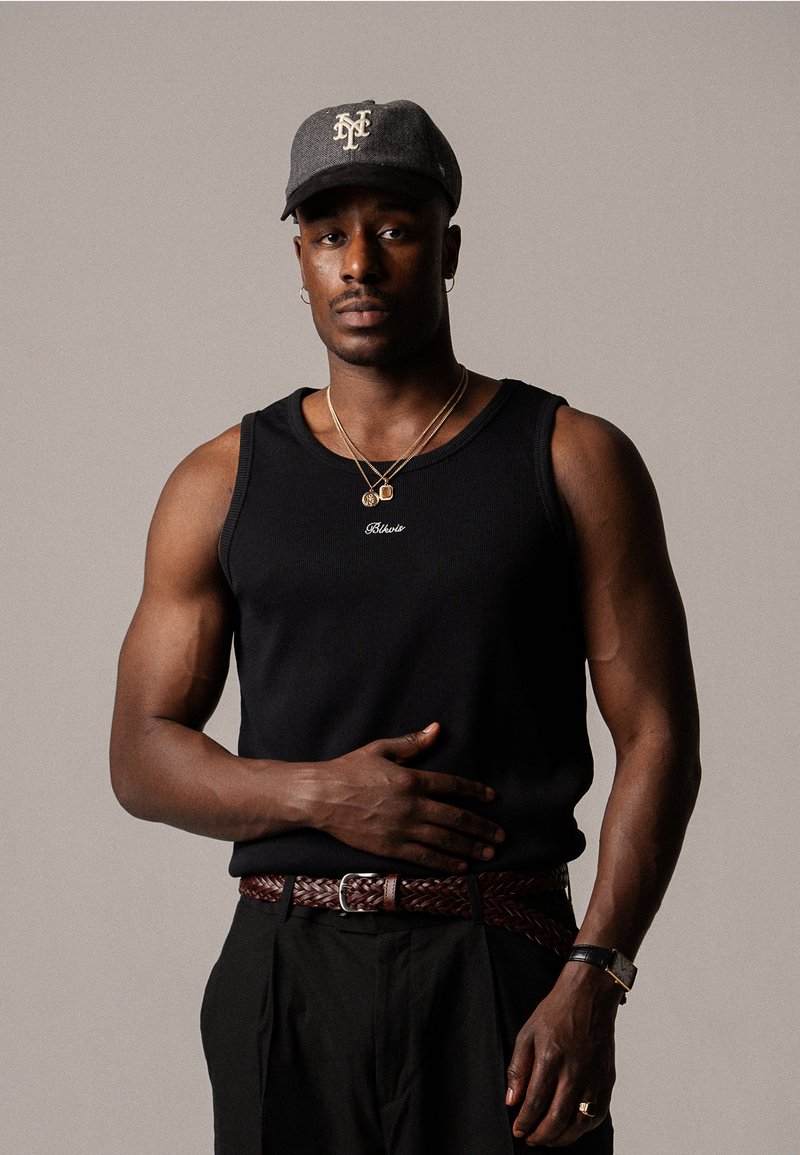 Débardeur noir avec bords côtelés, porté avec une ceinture tressée marron et des colliers en or superposés. Casquette grise avec des accents logo blancs.