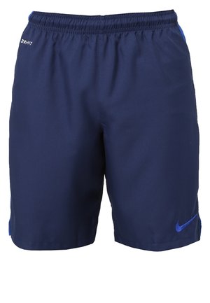 Homme portant un t-shirt blanc Nike, un short Nike bleu marine et des baskets blanches marchant vers l'avant sur un fond uni.