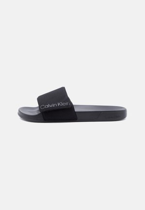 Zwarte Calvin Klein teenslipper met verstelbare band en logo op het voetbed en de zijkant van de zool, zijaanzicht op effen achtergrond.