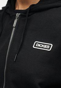 Κοντινό πλάνο μαύρου φούτερ με φερμουάρ, κορδόνια κουκούλας και λευκό κεντημένο λογότυπο «Dickies» στο αριστερό μέρος του στήθους.